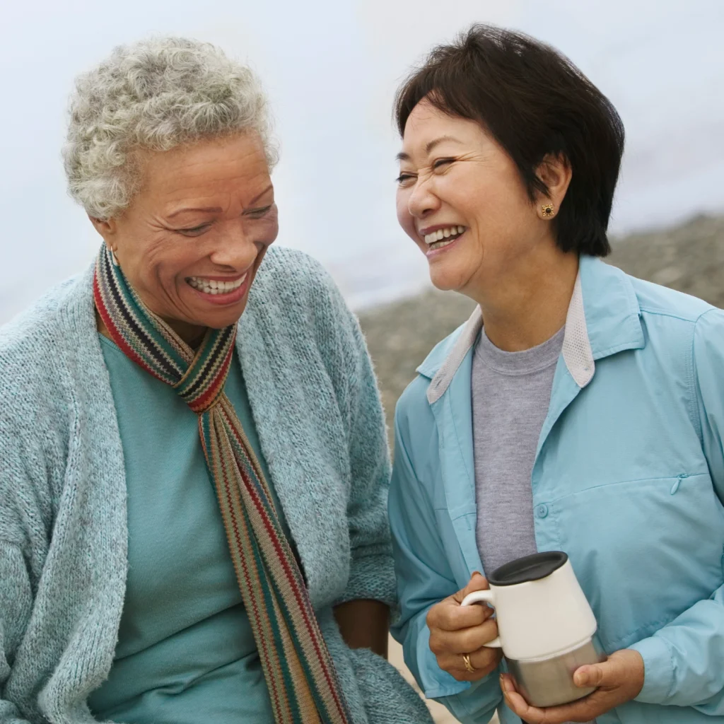 Two smiling older women chatting outdoors, one holding a travel mug